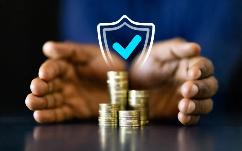 A pair of hands cupping stacks of coins with a glowing shield and checkmark icon above them, symbolising financial protection or insurance.