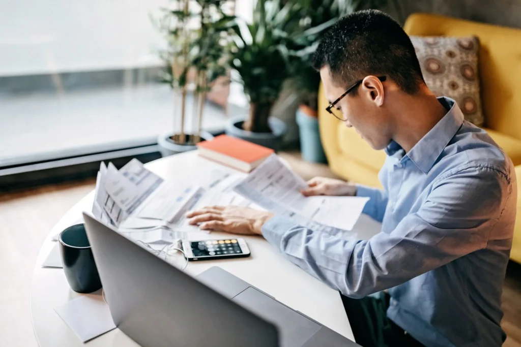 A man wearing glasses and a blue shirt sits at a white table, reviewing and organising several financial documents