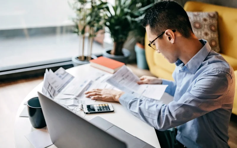 A man focused on reviewing several financial documents and receipts alongside a laptop and a smartphone.