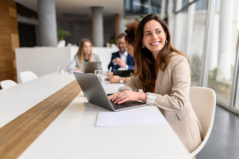 A smiling businesswoman in a tan blazer sits at a long white conference table