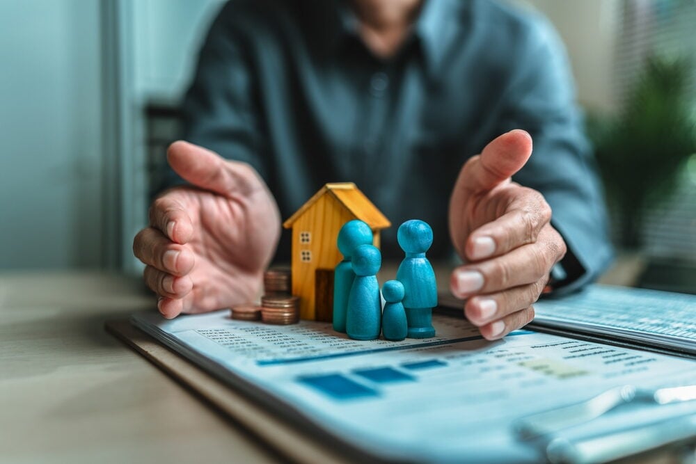 A person's hands protectively cupping a small wooden house model, a group of blue family figures, and stacks of coins on top of financial documents