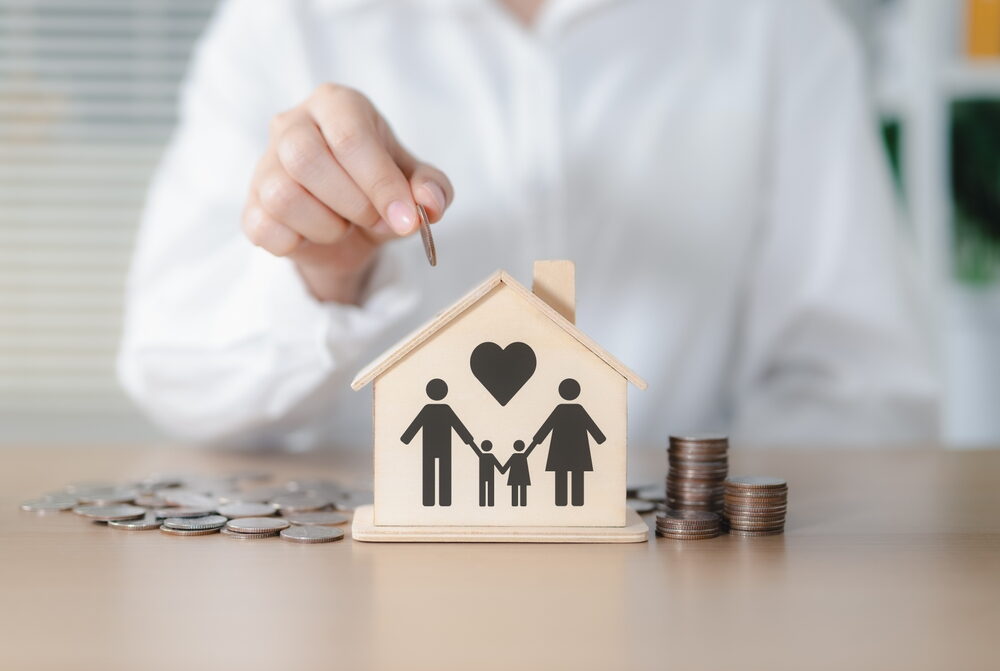 A person in a white shirt drops a coin into a wooden house-shaped piggy bank featuring a family icon and a heart