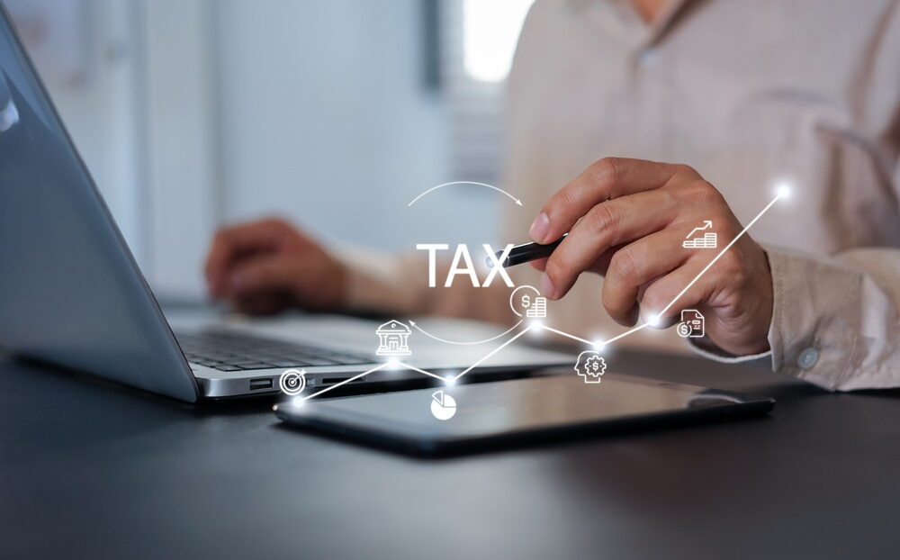 A person uses a stylus on a tablet while working on a laptop, featuring a digital "TAX" overlay